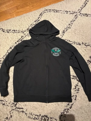 Svart Hammarby zip-hoodie - Snygg svart zip-hoodie från Hammarby med tryck på bröstet där det står 'Stockholm Hammarby South Side' och gröna vingar. Hoodien har dragkedja, två fickor fram och justerbar huva med snören. Perfekt för dig som vill representera ditt lag med stil.