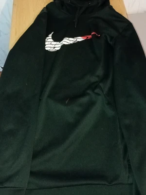 Svart Nike hoodie med rödvit logga - Svart hoodie från Nike med stor, stilren swoosh-logga på bröstet i vitt och rött. Loggan har texten 'NIKE' upprepat i ett mönster. Klassisk huva med dragsko och mjukt material som känns skönt mot huden. Perfekt för en sportig och avslappnad look.