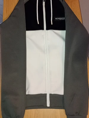 McKenzie hoodie i svart, vitt och grått - Snygg hoodie från McKenzie med dragkedja framtill. Färgblockad design i svart, vitt och grått. Huva med vita snören och logga på bröstet. Perfekt för en sportig och avslappnad stil.