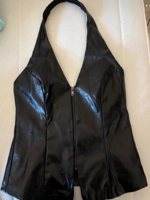 Svart halterneck topp i fejkskinn - Snygg svart halterneck topp i fejkskinn med dragkedja framtill. Toppen har en djup urringning och breda axelband som knäpps bakom nacken. Perfekt för en edgy och trendig look.