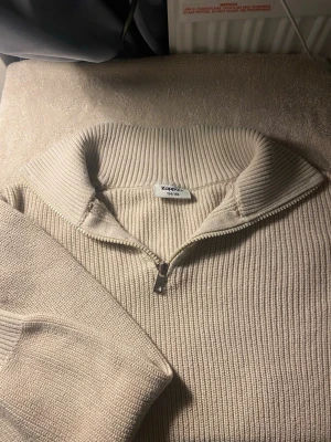 Beige stickad halvzip från Kappa - Säljer en beige stickad tröja från Kappa med halvzip och hög krage. Tröjan har ribbstickad struktur och långa ärmar, perfekt för lager på lager och en clean look.