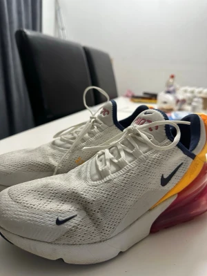 Nike Air Max 270 vita sneakers - Nike Air Max 270 sneakers i vitt meshmaterial med marinblå detaljer och en stor rosa Air Max-sula baktill. Skorna har gula och orange inslag vid hälen samt klassisk Nike-logga på sidan. Lätta och sportiga med modern design och bekväm passform.