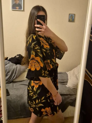 Svart blommig kortklänning med knyt - Snygg svart kortklänning med stora orange blommor och gröna blad. Klänningen har knyt i midjan och vida, korta ärmar. Perfekt för dig som vill sticka ut med ett färgstarkt mönster och bekväm passform. Passar xs/s. 