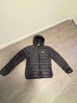 Svart dunjacka från EA7 Emporio Armani - Snygg svart dunjacka från EA7 Emporio Armani med huva och dragkedja framtill. Jackan har en quiltad design och logga på bröstet. Perfekt för kalla vinterdagar och riktigt lätt att bära. Klassisk passform och stilren look.