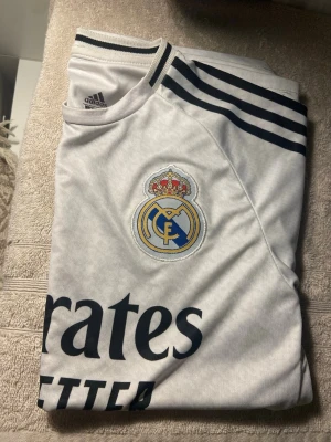 Real Madrid matchtröja Adidas vit - Säljer en vit Real Madrid matchtröja från Adidas med klubbmärke på bröstet och svarta detaljer. Tröjan har svarta ränder på axlarna, rund halsringning och trycket 'Emirates Fly Better' på framsidan. Perfekt för fotbollsfans!
