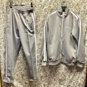 Grått tracksuit-set från Palm Angels - Snyggt grått tracksuit-set från Palm Angels med vita ränder längs ärmar och ben. Setet består av en zip-jacka med hög krage och matchande byxor med resår i midjan. Båda delarna har Palm Angels-logga tryckt i vitt. Perfekt för chill eller streetwear.