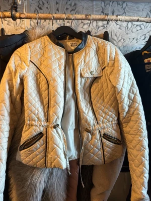 Beige quiltad jacka från Vero Moda - Snygg beige quiltad jacka från Vero Moda med dragkedja framtill, två fickor med dragkedjor och justerbar midja. Jackan har rundad krage och dekorativa sömmar som ger en cool look. Perfekt för dig som vill ha något stilrent och trendigt.