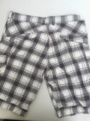 Rutig cargo shorts i bomull - Säljer ett par rutiga cargo shorts i vitt och grått med flera fickor och bälteshällor. Perfekta för sommaren och har en avslappnad passform. Tillverkade i mjuk bomull som känns skön mot huden.