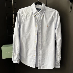 Ljusblå skjorta från Ralph Lauren - Klassisk ljusblå skjorta från Ralph Lauren med button-down krage och broderad logga på bröstet. Skjortan har långa ärmar, vita knappar och är tillverkad i mjuk bomull. Perfekt för en clean och stilren look.