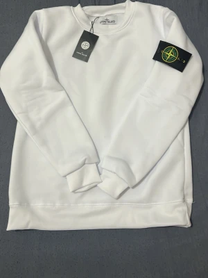 Vit sweatshirt från Stone Island - Snygg vit sweatshirt från Stone Island med klassisk rund halsringning och ribbade muddar. Tröjan har den ikoniska Stone Island-loggan på vänster ärm och är tillverkad i mjukt bomullsmaterial. Perfekt för en clean och stilren look.