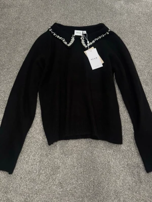 Svart stickad tröja med pärlkrage Vila - Supersnygg svart stickad tröja från Vila med lång ärm och fin krage dekorerad med vita pärlor. Tröjan är gjord i ett extremt mjukt och skönt material, sticks inte alls utan är jätte skön. Har ribbade muddar. Aldrig använd med prislapp kvar.