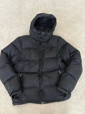 Svart pufferjacka från Polo Ralph Lauren - Snygg svart pufferjacka från Polo Ralph Lauren med huva och dragkedja framtill. Jackan har två fickor med dragkedja och den klassiska röda loggan broderad på bröstet. Perfekt för kalla vinterdagar och riktigt skön att bära.