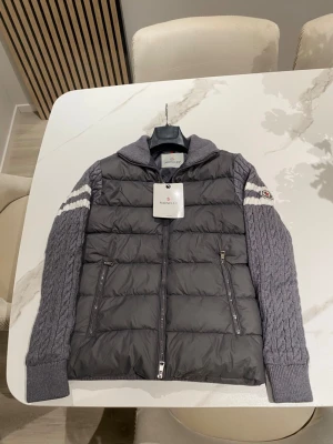 Grå Moncler cardiganjacka - Snygg grå cardiganjacka från Moncler med stickade ärmar och rygg i kabelstickat mönster. Framsidan är quiltad och vadderad med två dragkedjefickor. Ärmarna har vita ränder och ribbade muddar. Perfekt för dig som vill ha både stil och komfort.