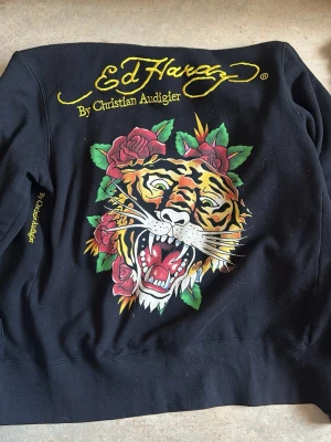  Rare Ed Hardy hoodie med tigertryck - Svart hoodie från Ed Hardy by Christian Audigier med stort färgglatt tigertryck och rosor på ryggen. Broderad gul logga upptill och texten 'Tattoo City' i gult på ena ärmen. Står EdHardy på framsidan med en ljusgrå tiger. Står ”by Christian Audigier” på sidan lodrätt vid midjan men täcks av ärmen såklart. Lite större i storlek, jag bär XS och denna är oversize, passar som oversize om man har XS, funkar bra med S och M och förmodligen tight om man har L