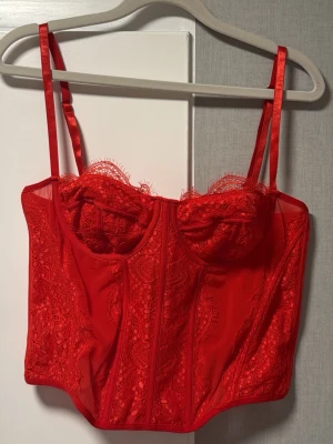 Röd spetskorsett med axelband - Snygg röd korsett med spetsdetaljer och justerbara axelband. Kuporna är dekorerade med spets och har lätt vaddering. Kroppen är i mesh med dekorativa sömmar och hakknäppning bak. Perfekt för att lyfta vilken outfit som helst. Använd 1 gång, lappen är tyvärr bortklippt men storleken är L