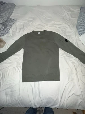 Grå sweatshirt från C.P. Company - Säljer en stilren grå sweatshirt från C.P. Company med rund hals och lång ärm. Tröjan har en ikonisk svart linsdetalj på ena ärmen och är tillverkad i mjuk bomull. Perfekt för en clean och avslappnad look.