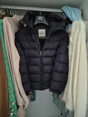 Svart dunjacka från Moncler - Snygg mörkblå dunjacka från Moncler med huva och dragkedja framtill. Jackan har quiltad design, Moncler-logga på ärmen och praktiska fickor. Perfekt för kalla vinterdagar och riktigt varm tack vare dunfyllningen. Några frågor skriv till mig!