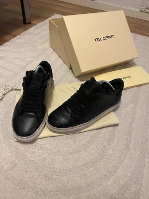 Svarta Axel Arigato sneakers i läder - Stilrena svarta sneakers från Axel Arigato i slät läder med svart snörning och vit sula. Klassisk låg modell med minimalistisk design och diskret logga på sidan. Perfekta för dig som gillar en clean och modern look.