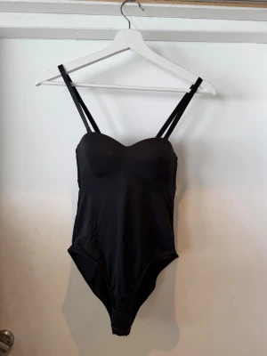 Svart body med justerbara axelband - Snygg svart body med dubbla, justerbara axelband och vadderade kupor. Bodyn har en smickrande sweetheart-ringning och knäppning i grenen med hyskor. Tillverkad i ett stretchigt, mjukt material som sitter bekvämt mot kroppen.
