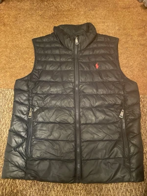 Svart dunväst från Ralph Lauren - Svart dunväst från Ralph Lauren med röd broderad logga på bröstet. Västen har två dragkedjeförsedda fickor och hög krage. Snygg, quiltad design som ger en sportig och stilren look. Perfekt att slänga över en hoodie eller tröja. Färgen är mörk mörk blå. OBS!!! Runt Large eller M i barnstorlek!!!