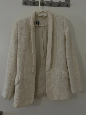 Krämvit bouclé kavaj från H&M - Stilren krämvit kavaj i bouclé från H&M med öppen front och två dekorativa fickor. Kavajen har en rak passform, helfodrad insida och fransade detaljer vid fickorna. Perfekt för att lyfta vilken outfit som helst med en chic touch.