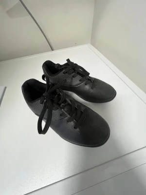 Svarta fotbollsskor med snörning strl. 31 - Säljer ett par svarta fotbollsskor med snörning och låg profil. Skorna har ett diskret rutmönster på sidorna och en logga i grått. Ovandelen är tillverkad i syntetmaterial som ger bra bollkontroll. Perfekta för dig som vill ha stilrena och funktionella skor på planen. Finns lite gräs kvar under skon men det påverkar inte funktionen.