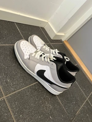 Nike vita och grå sneakers - Snygga sneakers från Nike i vitt och ljusgrått med svart swoosh och detaljer. Skorna har lågt skaft, klassisk snörning och perforerad tå för ventilation. Yttermaterialet är en mix av läder och syntet, och sulan är i gummi för bra grepp.