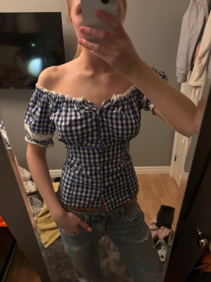 Blåvit rutig offshoulder blus - Säljer en blåvit rutig blus med offshoulder och korta puffärmar. Blusen har knappar framtill och spetsdetaljer vid ärmslut och halsringning. 