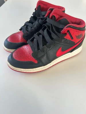 Nike Air Jordan 1 svart/röd sneakers - Säljer ett par klassiska Nike Air Jordan 1 sneakers i svart och rött läder med ikonisk vinge-logga på sidan och röd swoosh. Skorna har svart snörning, vit mellansula och röd yttersula. Perfekta för dig som gillar streetwear och retrostil.