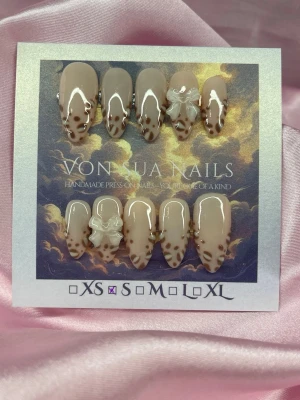 SoftLeo (S);Medium Almond - Handgjorda press-on naglar i nude och brunt med leopardmönster, dekorerade med 3D-vit rosett. Naglarna har en medium längd och har en almondform och en glansig finish. Perfekt för dig som vill ha en unik och trendig look.S: 15 mm, 11,5 mm, 12 mm, 11 mm, 9 mm 