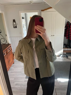 Grön jacka/overshirt - Militärgrön jacka/overshirt från Crocker. Rak passform och två stora bröstfickor. Jackan har klassisk krage, axelklaffar och knäppning framtill. Tillverkad i bomull. Helt oanvänd.