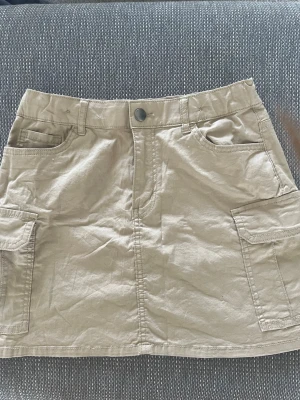 Beige cargoskort med fickor - Snygg beige cargoskort med flera praktiska fickor fram och på sidorna. Kjolen har bälteshällor, knappstängning och dragkedja. Tillverkad i ett mjukt bomullsmaterial med en avslappnad passform och casual look. Aldrig använd, prislappen kvar!🫰 Pris går att diskutera