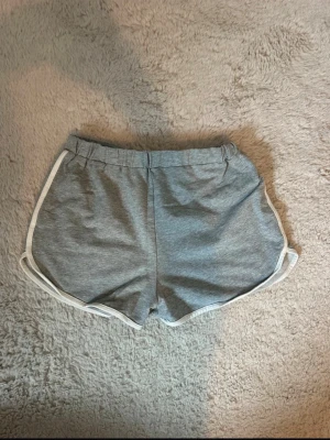 Grå shorts från SHEIN med vit kant - Grå shorts från SHEIN med elastisk midja och vita kanter längs sidorna. Klassisk sportig modell i mjukt bomullsmaterial, perfekta för sommardagar eller träning. Enkel och stilren design som är lätt att matcha.