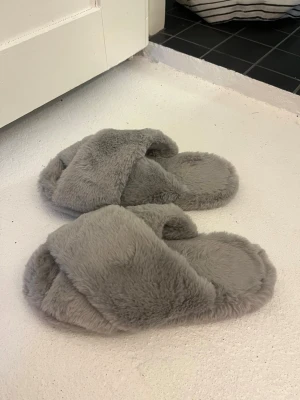 Grå fluffiga tofflor med korsad rem - Mysiga grå tofflor med korsad rem över foten och mjuk, fluffig yta. Perfekta för att hålla fötterna varma hemma. Tofflorna har öppen tå och är tillverkade i syntetiskt material som känns riktigt gosigt.