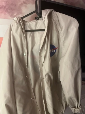 Beige NASA vindjacka med huva - Snygg beige vindjacka med NASA-logga på bröstet. Jackan har huva med dragsko, tryckknappar framtill och elastiska muddar vid ärmsluten. Tillverkad i lätt och vindtätt material, perfekt för blåsiga dagar.