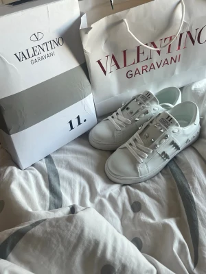 Vita Valentino Garavani sneakers - Snygga vita sneakers från Valentino Garavani med silverfärgade nitar och klassisk låg profil. Skorna har vita snören och är tillverkade i slätt läder med coola detaljer på sidan och tungan. Perfekta för dig som vill sticka ut med exklusiv streetstyle.