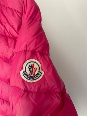 Rosa dunjacka från Moncler - Snygg rosa dunjacka från Moncler med quiltad design och dragkedja framtill. Jackan har två fickor med dragkedja och klassisk Moncler-logga på ärmen. Perfekt för kalla vinterdagar och riktigt lätt att bära. Materialet är glansigt och ger en modern look.