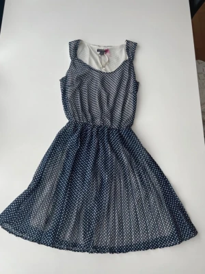 Marinblå prickig sommarklänning - Söt marinblå klänning med vita prickar och ärmlös design. Klänningen har en rundad halsringning och markerad midja som ger en fin siluett. Perfekt för varma dagar och enkel att styla med accessoarer.