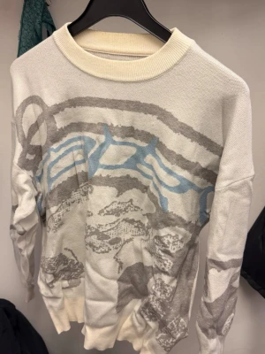 Vit stickad tröja med grafiskt mönster - Vit stickad tröja med rund hals och ribbade muddar. Tröjan har ett stort grafiskt mönster i grått och ljusblått över hela framsidan och ärmarna. Materialet ser ut att vara mjukt och bekvämt, perfekt för kyligare dagar.