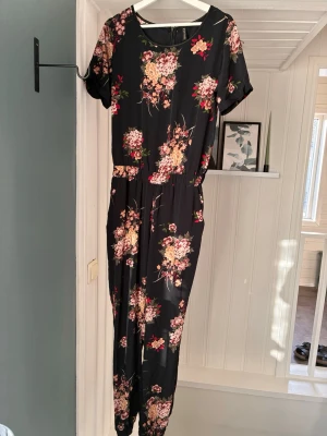 Svart blommig jumpsuit med kort ärm - Snygg svart jumpsuit med färgglatt blommönster i rosa, rött och beige. Modellen har korta ärmar, rund halsringning och resår i midjan för en skön passform. Lätt och mjukt material, perfekt för en trendig och avslappnad look.