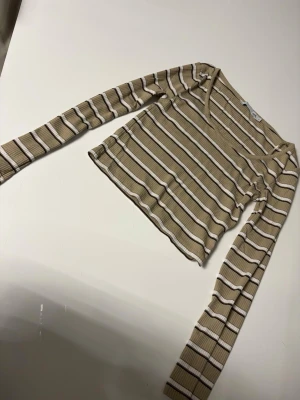 Beige och vit randig topp 157 - Säljer en ribbad långärmad topp från 157 i beige med vita och bruna horisontella ränder. Toppen har en djupare rundad urringning och är croppad i modellen. Perfekt till jeans eller kjol för en avslappnad look.