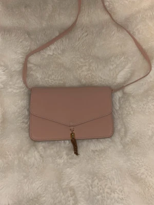 Väska - Söt ljusrosa handväska med axelrem och guldfärgad tofsdetalj framtill. Väskan har ett stilrent kuvertformat lock, spegel på insidan och guldfärgad kedja som kan användas för ett kortare band. Perfekt för att bära det viktigaste och ger en chic touch till din outfit. pris kan diskuteras. Tyvärr så hittar jag inte loggan för märket. 