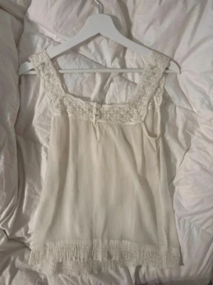 Vit spetsdetaljerad linnetopp - Söt vit linnetopp med breda axelband och fina spetsdetaljer runt ringningen. Toppen har små knappar framtill och smockad kant längst ner för extra stil. Perfekt för dig som gillar romantiska och feminina plagg.