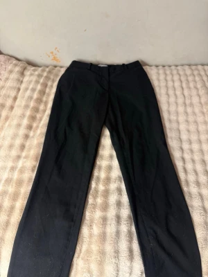 Svarta kostymbyxor från H&M - Snygga svarta kostymbyxor från H&M med klassisk rak passform och hällor för skärp. Byxorna har diskret dragkedja och knapp framtill. Perfekta för en stilren look och passar till många olika outfits.
