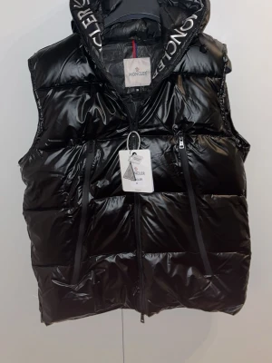 Svart dunväst från Moncler med huva - Snygg svart dunväst från Moncler med glansig finish och stor huva med Moncler-logga runt kanten. Västen har dragkedja framtill, två fickor med dragkedja och justerbara snören. Perfekt för lager-på-lager och streetwear-vibe.