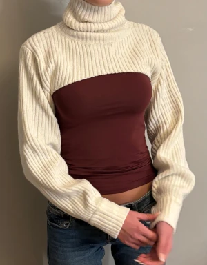 Vit ribbstickad bolero med polokrage - Vit ribbstickad bolero med hög polokrage och långa ärmar. Kort modell som slutar över bysten och ger en cool lager-på-lager-look. Perfekt att styla över linnen eller toppar för extra värme och stil.