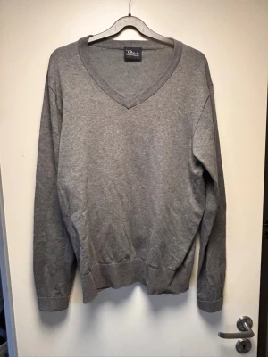 Grå V-ringad tröja från peak performance. - Klassisk grå V-ringad tröja från Peak performance. Tröjan har långa ärmar, ribbade muddar och en liten grön logga på ärmslutet. Perfekt basplagg i mjukt material som funkar till det mesta. Enkel och stilren design.