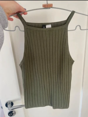 Olivgrön ribbad linnetopp - Säljer en olivgrön ribbad linnetopp med raka axelband och hög halsringning. Toppen är stickad och har en tight passform som sitter snyggt på kroppen. Perfekt till sommaren och enkel att matcha med jeans eller kjol.