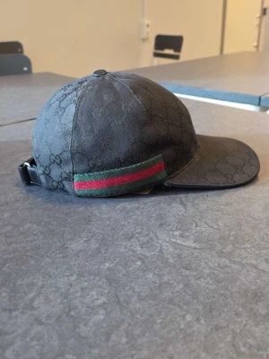 Svart Gucci keps med GG-mönster - Äkta  Snygg svart keps från Gucci med klassiskt GG-monogram över hela kepsen. ! BOX OCHH DUSTBAG FÖLJER MED! På sidan finns en grön och röd rand som ger den en ikonisk look. Justerbar rem bak för perfekt passform. Materialet är textil med exklusiv känsla.