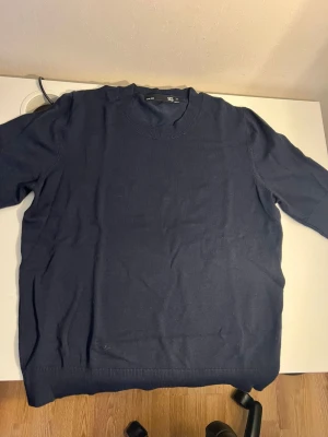 Mörkblå stickad tröja som har ett diskret hål - Mörkblå stickad tröja från Lager 157 med rund halsringning och ribbade muddar vid ärmslut och nederkant. Enkel och stilren design som passar till det mesta. Perfekt för lager på lager och funkar året runt.